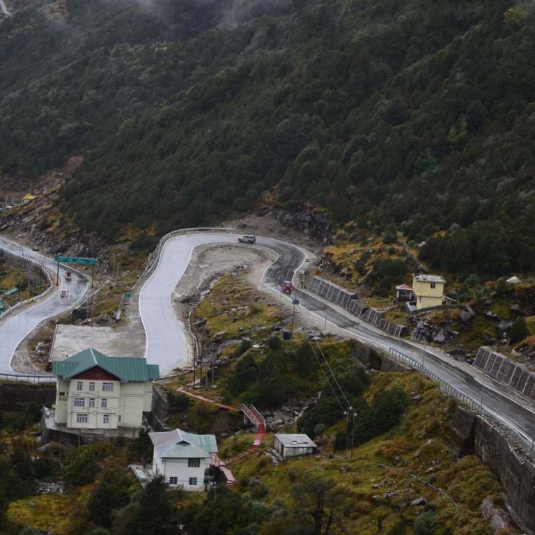 A winding road cutting through a mountain landscape with green trees. Two cars are on the road, and buildings are visible nearby.