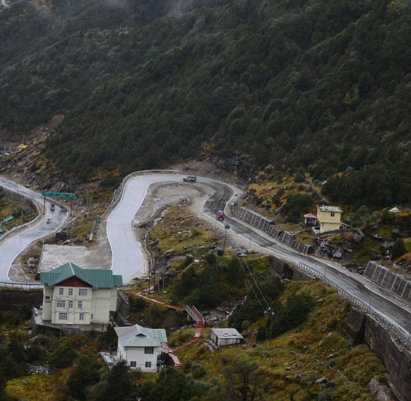 A winding road cutting through a mountain landscape with green trees. Two cars are on the road, and buildings are visible nearby.